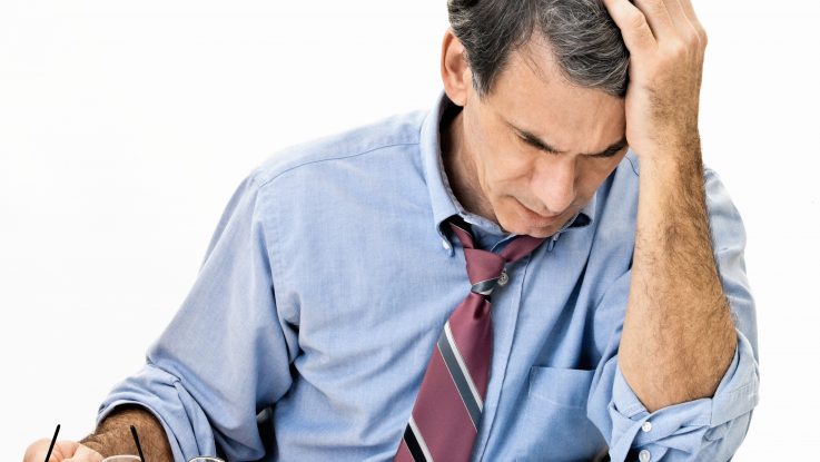 Man at desk in shirt and tie holding his head and worrying about money.